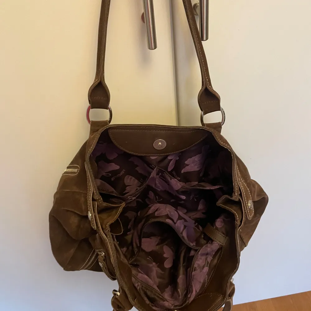 Snygg brun mockaväska från Longchamp med flera dragkedjor som kan användas för att göra väskan större och stiliga detaljer. Väskan har ett rymligt inre med ett lila fjärilsmönstrat foder. Den har en liten hake att fästa nycklar på, ett fack utanpå väskan och tre små fack inuti väskan. Perfekt för att bära med sig allt man behöver. Den har två handtag och en praktisk design. Självklart äkta. Man får plats med mappar från exacompta i den, så bra skolväska! Pris kan diskuteras!. Laukut & Käsilaukut.