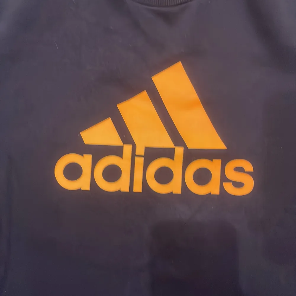 Säljer en svart tröja från Adidas med en stor orange logga på framsidan. Tröjan har långa ärmar och är perfekt för sportiga aktiviteter. Den är i storlek 164, vilket passar ungdomar i åldern 13-14 år.. Neuletakit & Villapaidat.