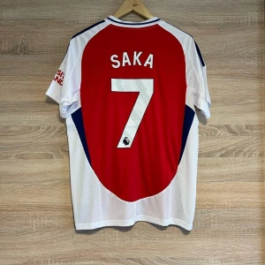 Röd och vit Arsenal fotbollströja XL - Säljer en snygg Arsenal fotbollströja från Adidas i rött och vitt med korta ärmar. Tröjan har spelarnamnet 'Saka' och nummer 7 på ryggen. Perfekt för alla Arsenal-fans! 🔴⚪