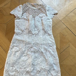 Vit spetsklänning från Ida Sjöstedt - Elegant vit spetsklänning från Ida Sjöstedt med korta ärmar och ett vackert prickigt mönster på överdelen. Klänningen har en fin spetsdetalj som ger en romantisk känsla. Perfekt för speciella tillfällen.
