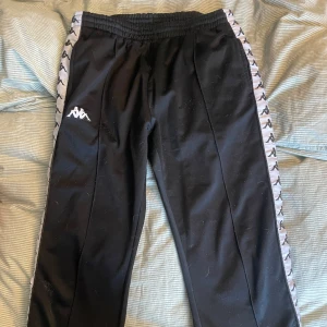 Svarta träningsbyxor från Kappa - Snygga svarta träningsbyxor från Kappa med det klassiska logotypbandet längs sidorna. Byxorna har en elastisk midja för extra komfort och är perfekta för en sportig look.
