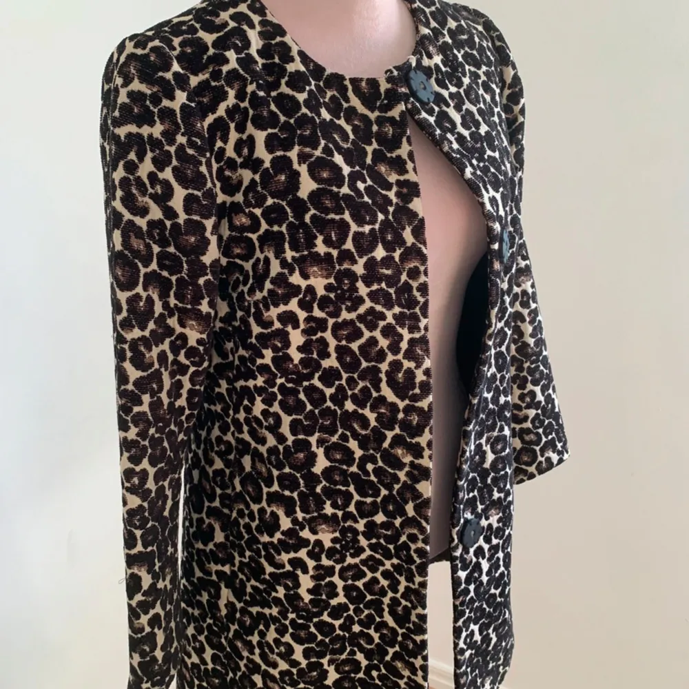 Snygg leopardmönstrad kappa med stora svarta knappar framtill. Den har en klassisk passform och är perfekt för att ge en trendig touch till din outfit. Kappan har långa ärmar och en rund halsringning.. Takit.