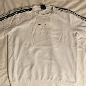 Vit sweatshirt från Champion - Säljer en vit sweatshirt från Champion med svarta detaljer längs ärmarna. Tröjan har Champion-loggan broderad på bröstet och längs ärmarna. Perfekt för en sportig look eller avslappnad stil.