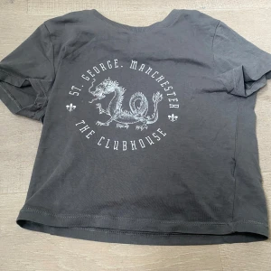 Grå t-shirt från H&M Divided - Säljer en grå t-shirt från H&M Divided med tryck av en drake och texten 'St. George, Manchester, The Clubhouse'. T-shirten är kortärmad och i bomull, perfekt för en avslappnad stil.