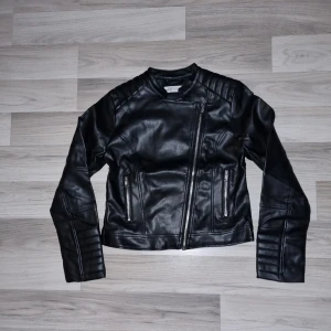 Svart jacka från H&M - Snygg svart jacka i skinnimitation från H&M med dragkedja och coola detaljer. Jackan har långa ärmar och två fickor med dragkedjor framtill. 