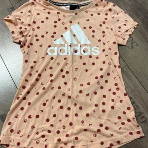 Persikofärgad t-shirt med prickar från Adidas - Snygg persikofärgad t-shirt från Adidas med röda prickar och det klassiska logotyptrycket i vitt på bröstet. Perfekt för en sportig look eller avslappnad stil. Kortärmad och tillverkad i mjukt material för extra komfort. Obs liten i storleken så även om det är en L skulle jag säga att det är en s/m!