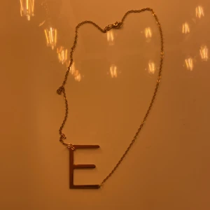 Guldhalsband med bokstaven E - Elegant guldhalsband med en stor bokstav E som hänge. Perfekt för att ge en personlig touch till din stil. Kedjan är tunn och smidig, vilket gör den bekväm att bära.