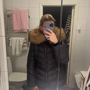 Svart dunjacka från Saki - Snygg svart dunjacka från Saki med en stor, med äkta minkpäls på huvan. Jackan har dragkedja och ett bälte runt midjan.
