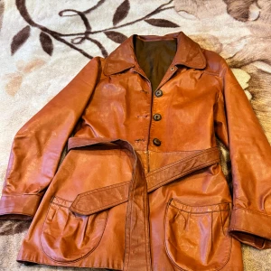 Brun skinnjacka med bälte - Snygg brun skinnjacka med knappar och bälte i midjan. Jackan har långa ärmar och stora fickor framtill. Perfekt för höst och vår.