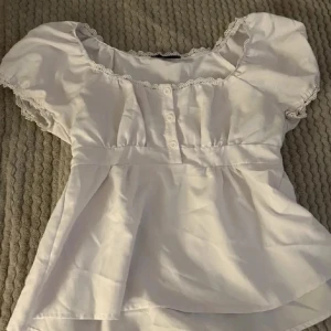 Jätte söt topp - Charmig vit blus med spetsdetaljer vid ärmslut och hals. Blusen har korta ärmar och knappar framtill, vilket ger en romantisk touch. Perfekt för en somrig look. 