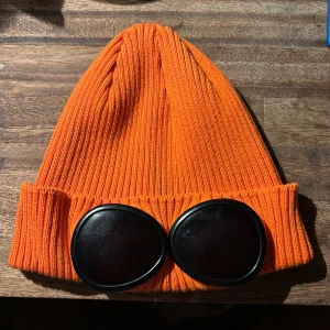 C.P company mössa - Snygg orange märkes mössa med ribbad design och coola glasögonliknande detaljer framtill. Perfekt för att sticka ut i mängden och hålla sig varm. Köptes på farfetch. Ny skick!