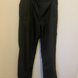 Svarta regnbyxor för barn/undom - Svarta byxor med högteknologisk väv i nylon och polyester. De har tejpade sömmar och är vattentäta med en vattenresistens på 5,000 mm. Bra andningsförmåga på 3,000 g/m²/24h. Perfekta för regniga dagar.