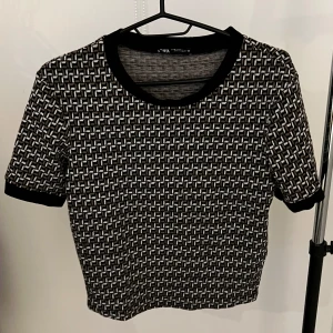 Mönstrad topp från Zara - Snygg mönstrad topp från Zara i svart och grått med korta ärmar. Toppen har ett geometriskt mönster och svarta kanter vid ärmslut och halsringning. Perfekt för en stilren look!