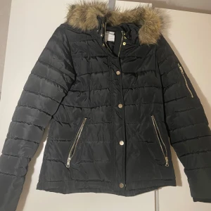 Svart dunjacka med fuskpäls - Snygg svart dunjacka med fuskpäls på huvan och dragkedjor i guld. Jackan har långa ärmar och stängs med både dragkedja och knappar. Perfekt för kyliga dagar.