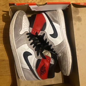 Nike Air Jordans i grått, vitt och rött - Säljer ett par Nike Air Jordans i en snygg kombination av grått, vitt och rött. Skorna har den klassiska höga modellen med svart snörning och Nike-logga. Perfekta för sneaker-entusiaster!