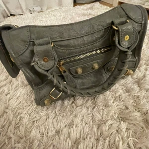 Grå axelväska med dragkedja - Snygg grå axelväska i skinn med detaljer som dragkedja och knappar. Väskan har en flätad handtag och guldfärgade metalldetaljer som ger en lyxig känsla.