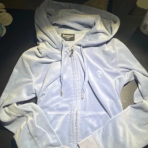 Ljusblå hoodie från Juicy Couture - Säljer en mjuk och stilren ljusblå hoodie från Juicy Couture. Den har dragkedja och en praktisk huva med snörning. Perfekt för en avslappnad look.