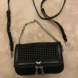 Svart axelväska med nitar från Zadig & Voltaire - Snygg svart axelväska från Zadig & Voltaire med silverfärgade nitar och kedjedetaljer. Väskan har en justerbar axelrem och dubbla dragkedjor framtill. Säljer för att den inte kommer till användning.☺️