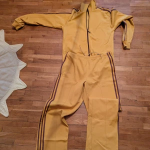 Adidas tracksuit orginal Vintage  - Snygg gul träningsoverall med bruna ränder längs ärmar och ben. Jackan har dragkedja och hög krage, medan byxorna har en bekväm passform. Perfekt för en retro look! Visst slitage längst ner.annars bra skick 