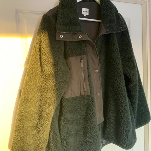 Grön teddyjacka från Indiska - Mysig grön teddyjacka från Indiska med dragkedja och knappar framtill. Jackan har en hög krage perfekt för kyliga dagar. Den har en praktisk bröstficka med dragkedja. Väldigt sparsamt använd.