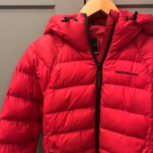 Röd dunjacka från Peak Performance - Snygg och varm röd dunjacka från Peak Performance med huva och dragkedja. Perfekt för kyliga dagar. 