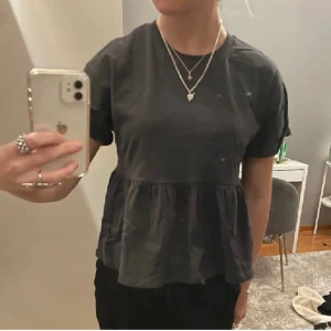 Mörkgrå blus med volangdetalj - Säljer en stilren mörkgrå blus med korta ärmar och volangdetalj nedtill. Säljer då den inte kommer till användning. Lånade bilder🤗