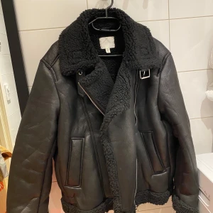 Svart jacka från H&M - Säljer en svart jacka från H&M med en mysig teddyfoder och bred krage. Jackan har dragkedja och två fickor framtill. Perfekt för kyliga dagar.