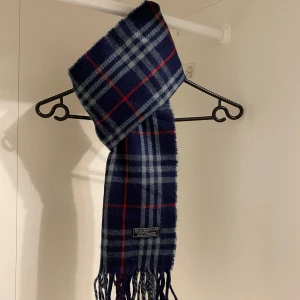 Burberry - Säljer nu denna Snygga halsduk från Burberry i blått.  Den har klassiska fransar i ändarna och är perfekt för att hålla dig varm under kyliga dagar. Ett stilrent val för både höst och vinter. Den är gjord i mjukt lammull. Hör av er vid funderingar☺️