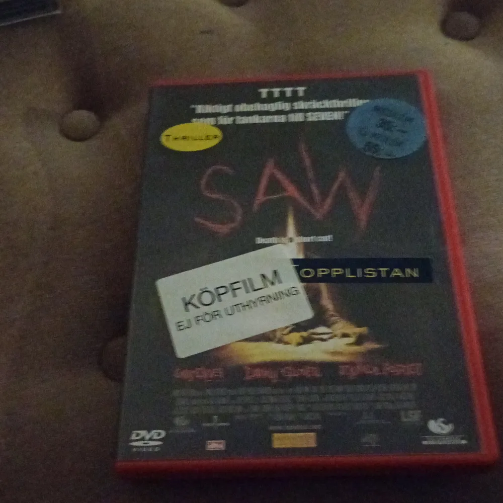 DVD-film av skräckfilmen 'Saw' i ett rött fodral. Omslaget har titeln i röd text och flera klistermärken, inklusive 'Köpfilm' och 'Topp 10'. Perfekt för skräckfilmsentusiaster som vill ha en klassiker i samlingen.. Muu.
