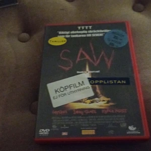 Saw DVD - DVD-film av skräckfilmen 'Saw' i ett rött fodral. Omslaget har titeln i röd text och flera klistermärken, inklusive 'Köpfilm' och 'Topp 10'. Perfekt för skräckfilmsentusiaster som vill ha en klassiker i samlingen.