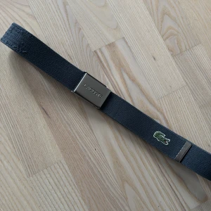Gråblått bälte från Lacoste - Snyggt gråblått bälte från Lacoste med silverfärgat spänne och den klassiska krokodilloggan. Perfekt för att ge en stilren touch till din outfit.
