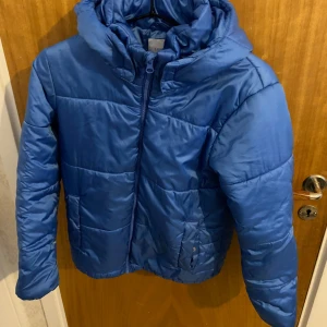 Blå pufferjacka från Lindex Kids - Säljer en blå pufferjacka från Lindex Kids. Jackan har en dragkedja framtill och en mysig huva. Perfekt för kyliga dagar med sin quiltade design. Har haft det länge den kan vara bra julklapp för barn mellan 7-10