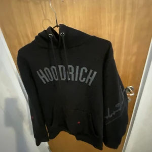 Svart hoodie från Hoodrich - Säljer en svart hoodie från Hoodrich med tryck på både fram- och baksidan. Den har en klassisk känguruficka och justerbar huva med dragsko. Perfekt för en avslappnad stil. Men  svarta färgen har blivit blekare 