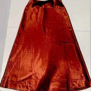 Rödvinfärgad långkjol i satin  - Elegant rödvinfärgad långkjol i satin med en mjuk och glansig yta. Kjolen har en klassisk A-linjeform som ger en vacker silhuett. Perfekt för att ge en touch av lyx till din garderob. Velvet tyg insidan. Oanvänd. Free size.