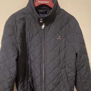 Mörkblå quiltad jacka från Gant - Snygg mörkblå quiltad jacka från Gant med dragkedja och knappdetaljer vid kragen. Perfekt för kyligare dagar med sin stilrena design och bekväma passform. Är i mycket fint skick och knappt använd, perfekt för kommande vår, Storlek : S