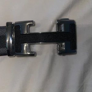 Svart och grått bälte med rutigt mönster - Snyggt bälte med ett stilrent rutigt mönster i svart och grått. Bältet har ett elegant spänne i silver och svart som ger en modern touch. Perfekt för att komplettera din outfit.