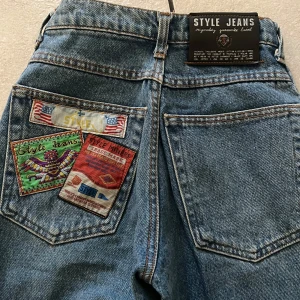 Blå jeans med patchar från Style Jeans - Säljer ett par coola blå jeans från Style Jeans med unika patchar på bakfickorna. Jeansen har en klassisk femficksdesign och knappgylf. Perfekta för en avslappnad stil.