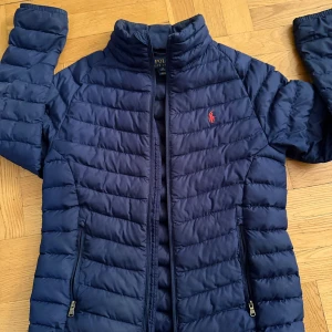 Blå pufferjacka från Ralph Lauren - Säljer en blå pufferjacka från Ralph Lauren med en dragkedja som är i  behov av kärlek det är även andra slitningar som kan behöva sys ihop.Det står att jackan är storlek M men passar Xs/S.