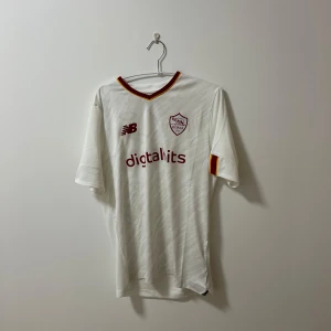 Roma fotbollströja  - Snygg vit fotbollströja från New Balance med röda detaljer och klubbmärke på bröstet. Tröjan har korta ärmar och en V-ringad hals med röd och gul kant. Perfekt för fotbollsfans!