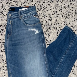Blå jeans från Replay - Snygga blå jeans från Replay med slitna detaljer och klassisk femficksdesign. De har en normal passform och är perfekta för en avslappnad stil. Märkeslogga på baksidan av midjan. (Använda 1 gång)