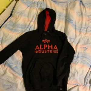 Svart hoodie från Alpha Industries - Säljer en svart hoodie från Alpha Industries med röd text och logga på framsidan. Hoodien har en röd insida i huvan och dragsko. Perfekt för en casual look.