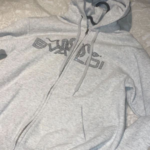 Grå hoodie med dragkedja - Snygg grå hoodie med dragkedja och tryck framtill. Perfekt för en avslappnad stil. Den har en justerbar huva med snören och är tillverkad i ett mjukt material. Ifrån bvaldi 