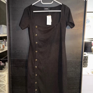 Svart ribbad klänning med knappar - Snygg svart ribbad klänning från Cider med knappar längs sidan. Klänningen har korta ärmar och en fyrkantig halsringning. Perfekt för en stilren look.