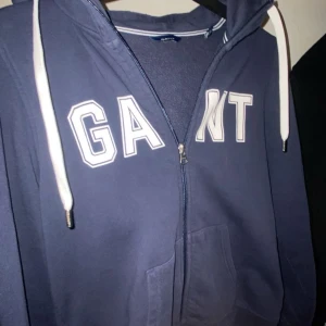 Mörkblå hoodie från Gant - Säljer en snygg mörkblå hoodie från Gant med dragkedja och vit text på bröstet. Den har en klassisk design med justerbar huva och ribbade muddar. Perfekt för en avslappnad stil. Storlek xs skulle nog rekommendera den till någon runt 160-165