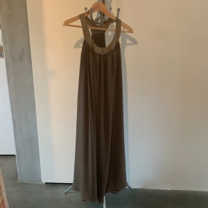 Brun långklänning från Monday  - Elegant brun långklänning från Monday med en lös passform. Klänningen har en ärmlös design och ett stilrent snitt som ger en flytande siluett. Perfekt för en stilren look.