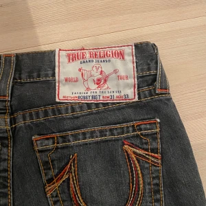 Svarta  True Religion jeans - Snygga svarta jeans från True Religion med klassisk design och orange sömmar. De har en bootcut-stil och är perfekta för en avslappnad look. Jeansen har en högkvalitativ denimkänsla och en ikonisk logga på baksidan. Skriv för frågor och mått osv