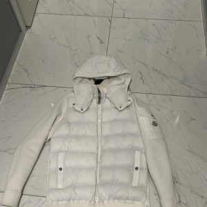 Vit cardigan från Moncler - Säljer en snygg vit cardigan från Moncler med avtagbar huva och dragkedja framtill. Jackan har långa ärmar och ribbade muddar. Perfekt för kyliga dagar med stil. 🏔️