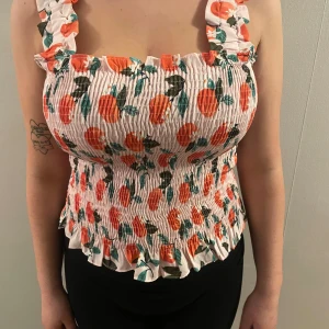 Blommig smocktopp från Pieces - Säljer en söt blommig smocktopp från Pieces i vitt med orange blommönster. Toppen har volangdetaljer och breda axelband. Perfekt för en somrig look! 🌸