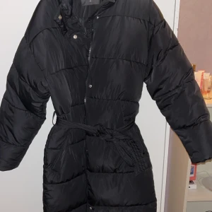 Svart dunjacka med bälte - Snygg svart dunjacka med bälte i midjan för en figursydd look. Jackan har långa ärmar och stängs med dragkedja och knappar. Perfekt för kalla dagar.