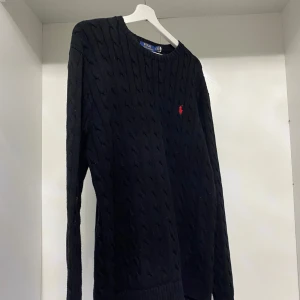 Svart stickad tröja från Polo Ralph Lauren - Säljer en stilren svart stickad tröja från Polo Ralph Lauren med kabelstickat mönster. Tröjan har långa ärmar och en liten röd logotyp på bröstet. Perfekt för en klassisk och tidlös look. Passar fint till jeans! Storlek L, men passar som M också.